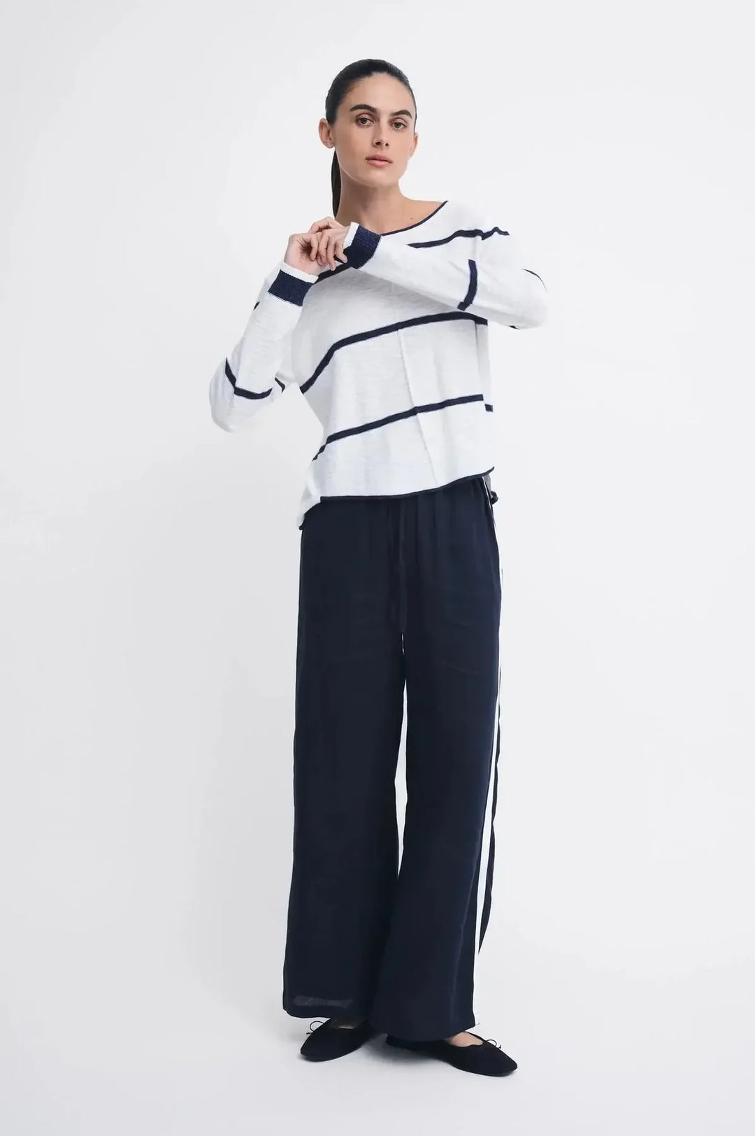A woman modeling a white knit sweater with navy blue horizontal stripes and matching navy wide-leg trousers, standing against a plain light gray studio background.
