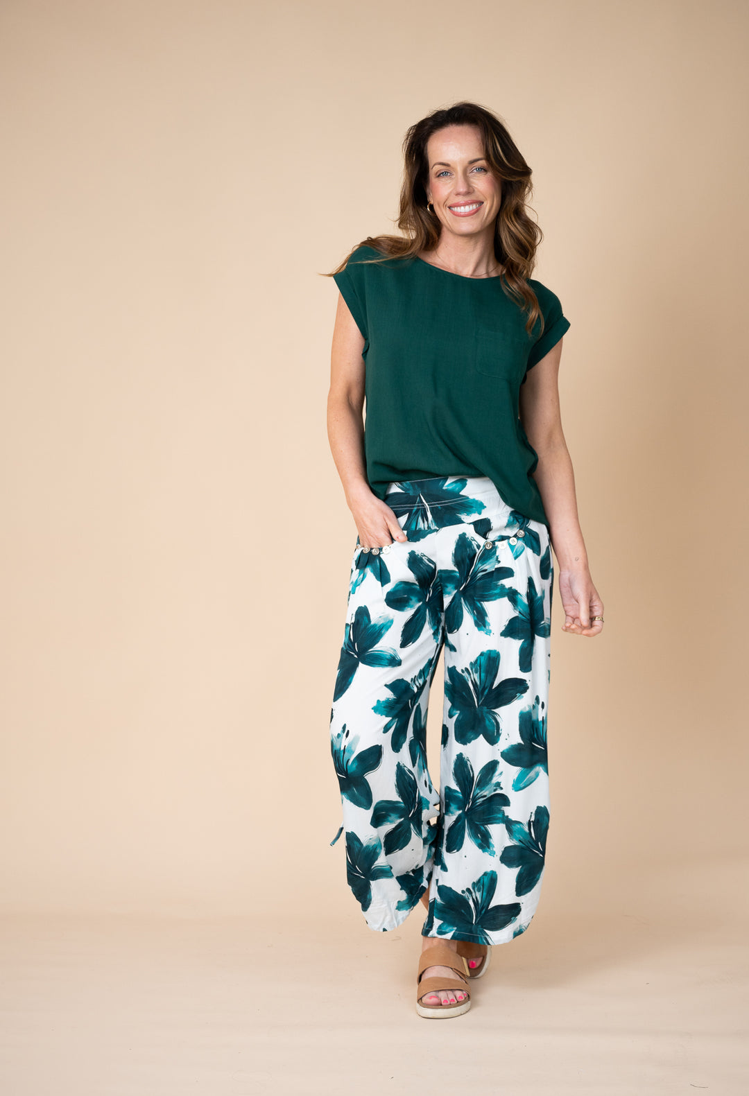 A woman modeling a dark green short-sleeve top and white wide-leg pants with a dark green tropical floral print, standing against a solid beige studio background.