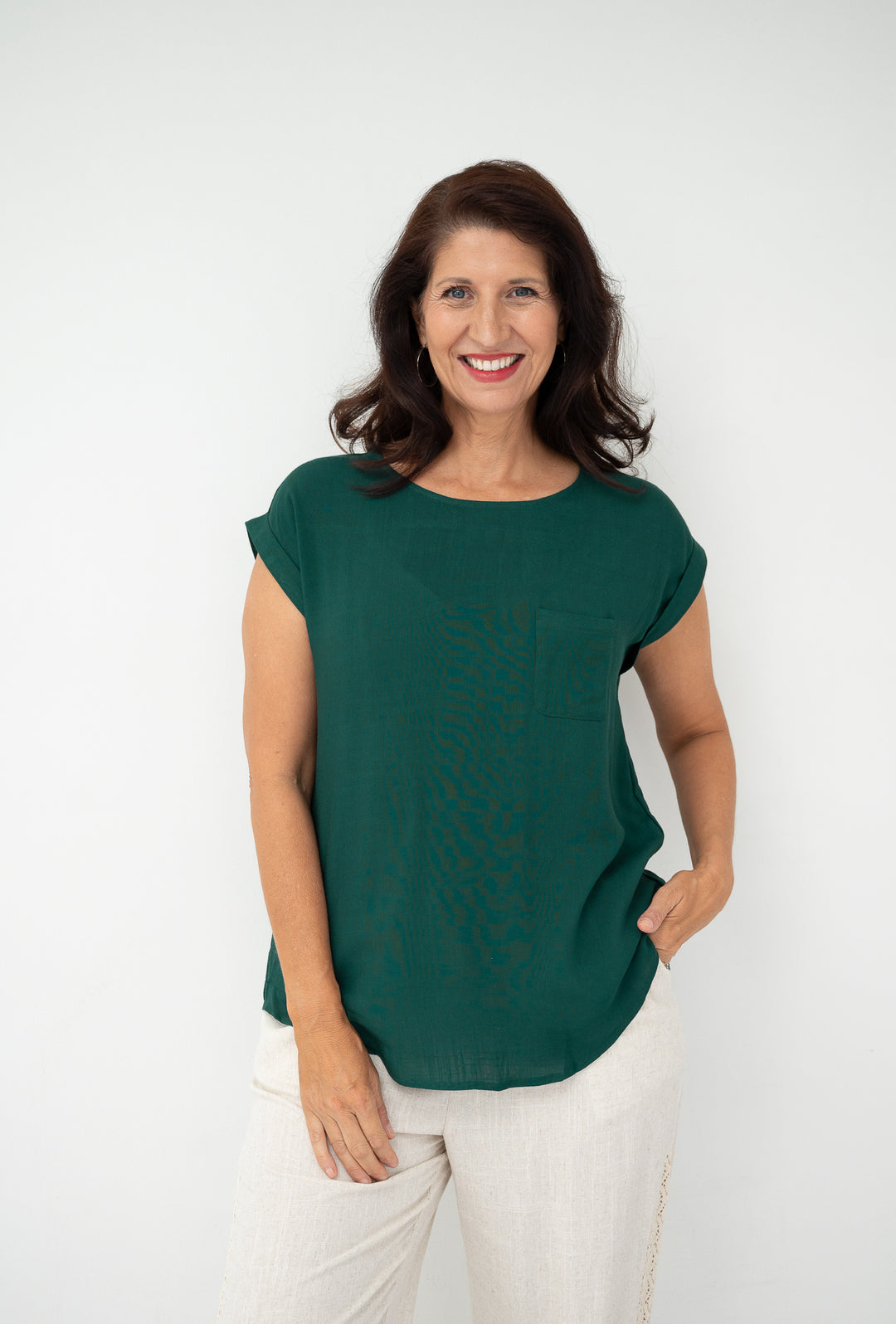 A smiling woman with dark brown hair wearing a dark green short-sleeve top and white pants, standing against a plain white studio background.