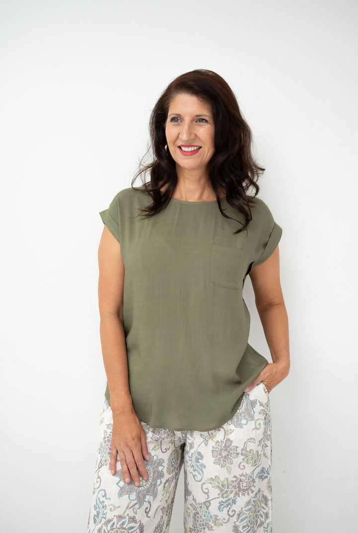 A woman with dark hair smiling, wearing a short-sleeve olive green top and white floral pants, standing against a plain white background.