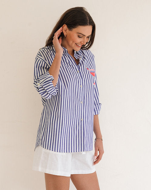 A woman wearing a blue and white vertically striped button-down shirt with rolled-up sleeves and white shorts, standing against a plain off-white background.