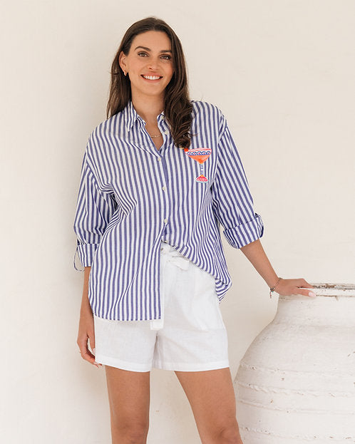 A woman wearing a blue and white vertically striped button-down shirt with a small orange and red embroidered pocket, paired with white shorts, standing next to a large white ceramic vase against a plain off-white wall.