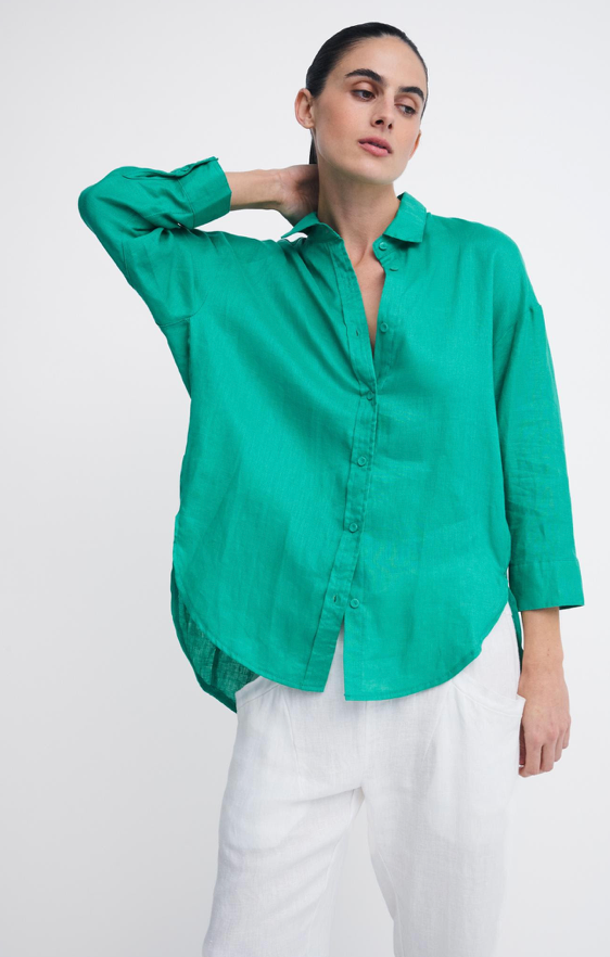 A woman modeling a bright green linen button-up shirt with white pants, posing against a light gray studio background.