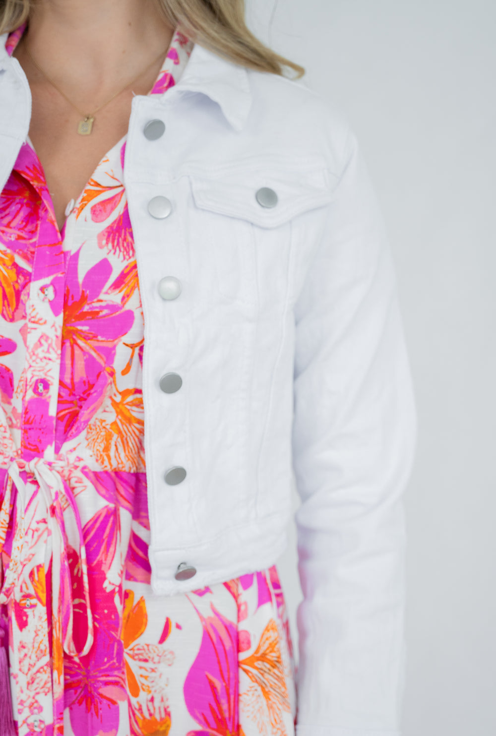 A close-up of a person wearing a white denim jacket over a bright pink, orange, and white floral dress against a light gray background.