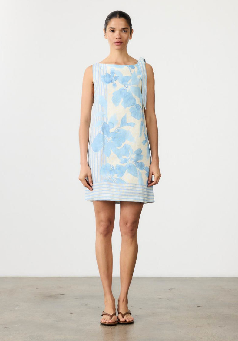 A woman stands facing forward in a sleeveless, knee-length dress with a light blue floral print on a white base, paired with brown sandals, against a plain white studio wall.