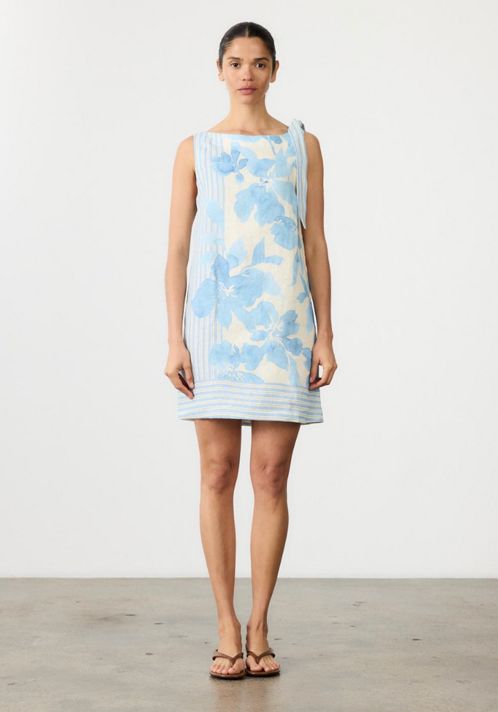 A woman stands facing forward in a sleeveless, knee-length dress with a light blue floral print on a white base, paired with brown sandals, against a plain white studio wall.