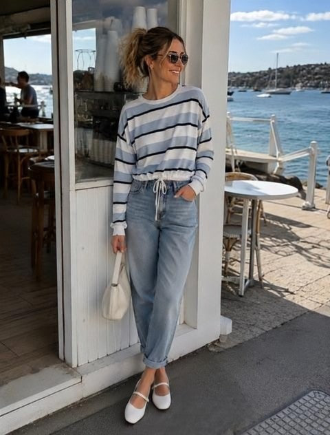 A woman stands on a waterfront promenade wearing a blue and white striped long-sleeve crop top, light-wash blue jeans, white Mary Jane shoes, and holding a small white handbag.