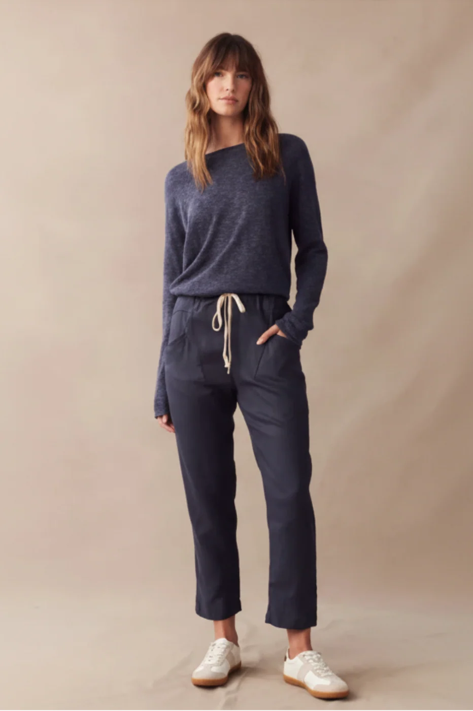A woman stands against a plain beige studio backdrop wearing a navy knit sweater, navy drawstring pants, and white sneakers.