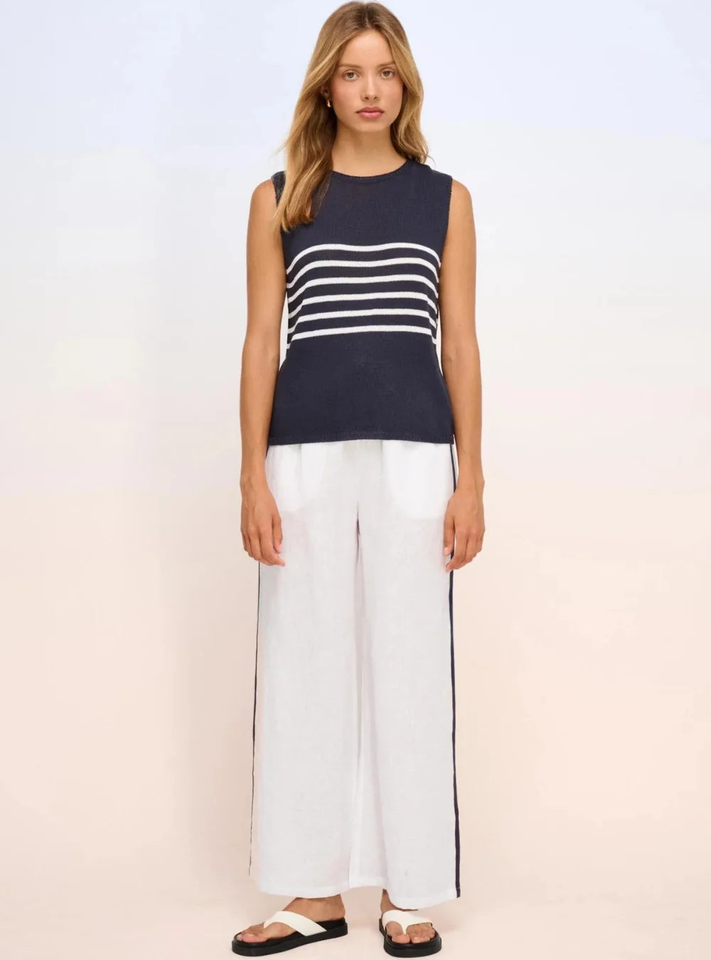 A woman modeling a sleeveless knit top with navy blue and white horizontal stripes, paired with white wide-leg pants and black-and-white sandals, against a light studio background.