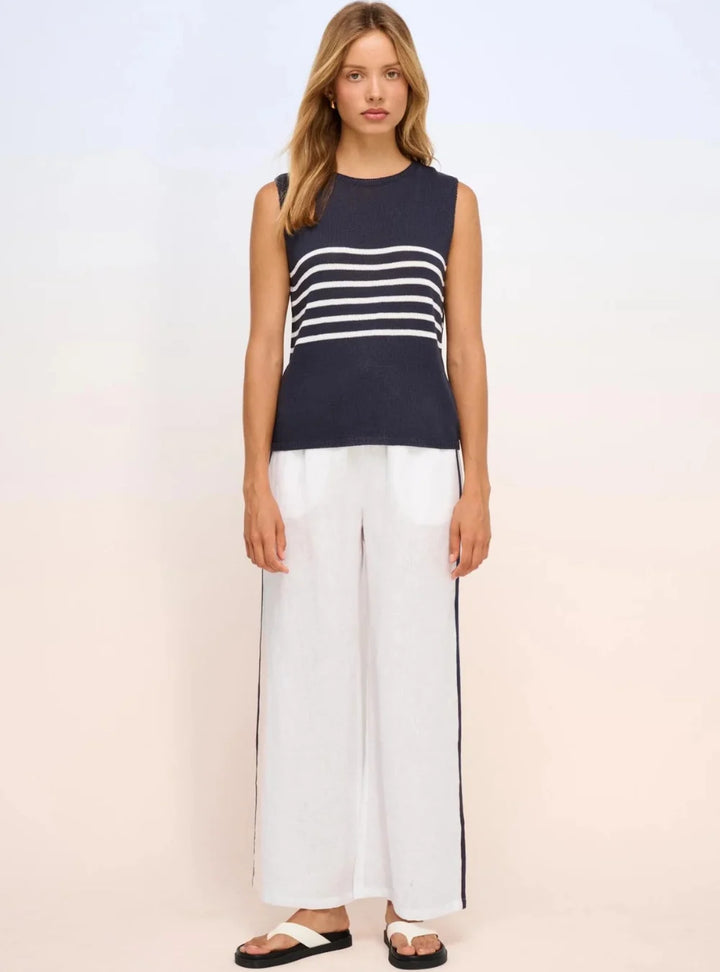 A woman modeling a sleeveless knit top with navy blue and white horizontal stripes, paired with white wide-leg pants and black-and-white sandals, against a light studio background.