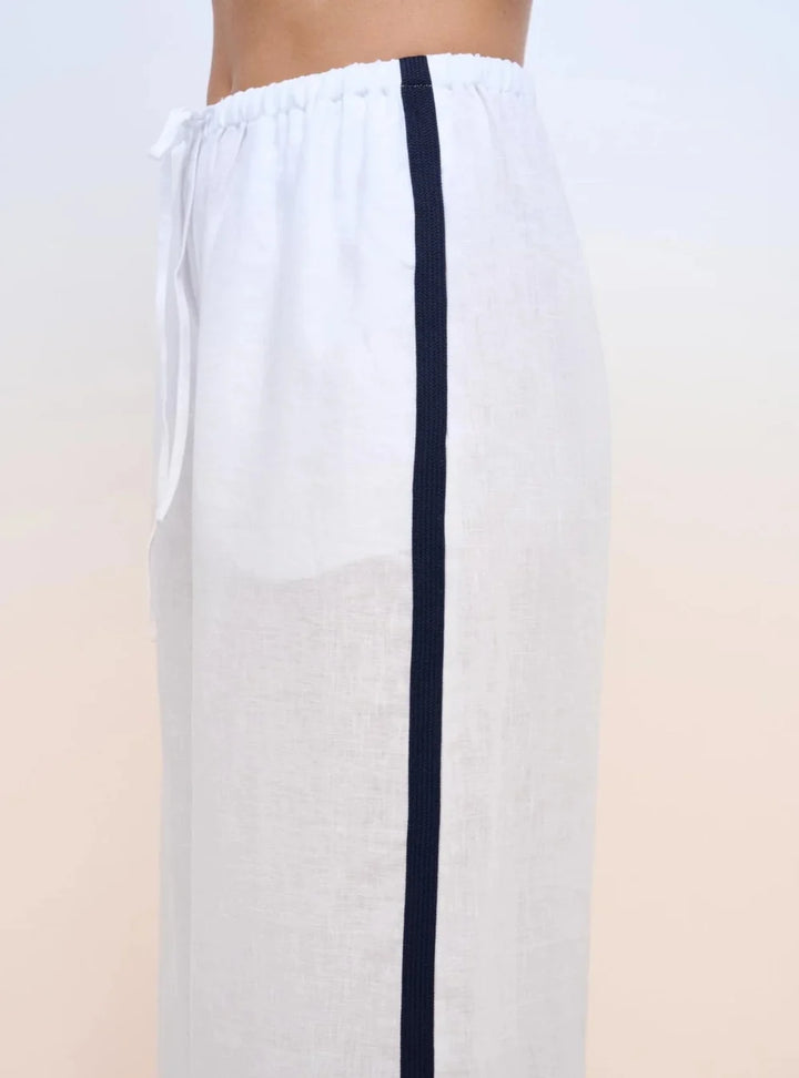 White linen drawstring pants with a vertical navy blue stripe down the side, shown on a model cropped from the waist down against a light beige gradient background.