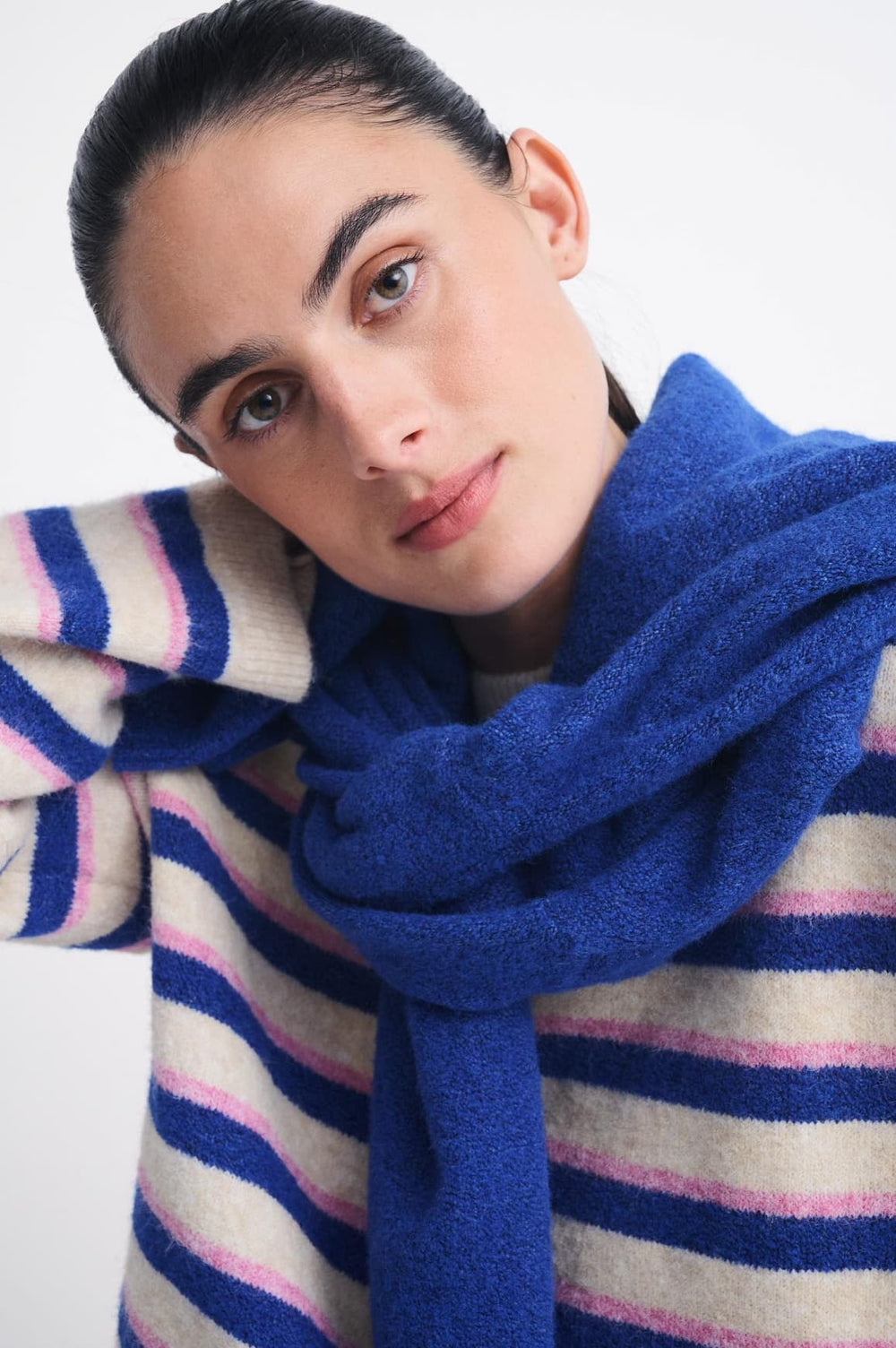 A close-up of a woman wearing a thick, vibrant royal blue knit scarf and a striped sweater with navy, pink, and cream horizontal stripes, set against a plain light gray background.