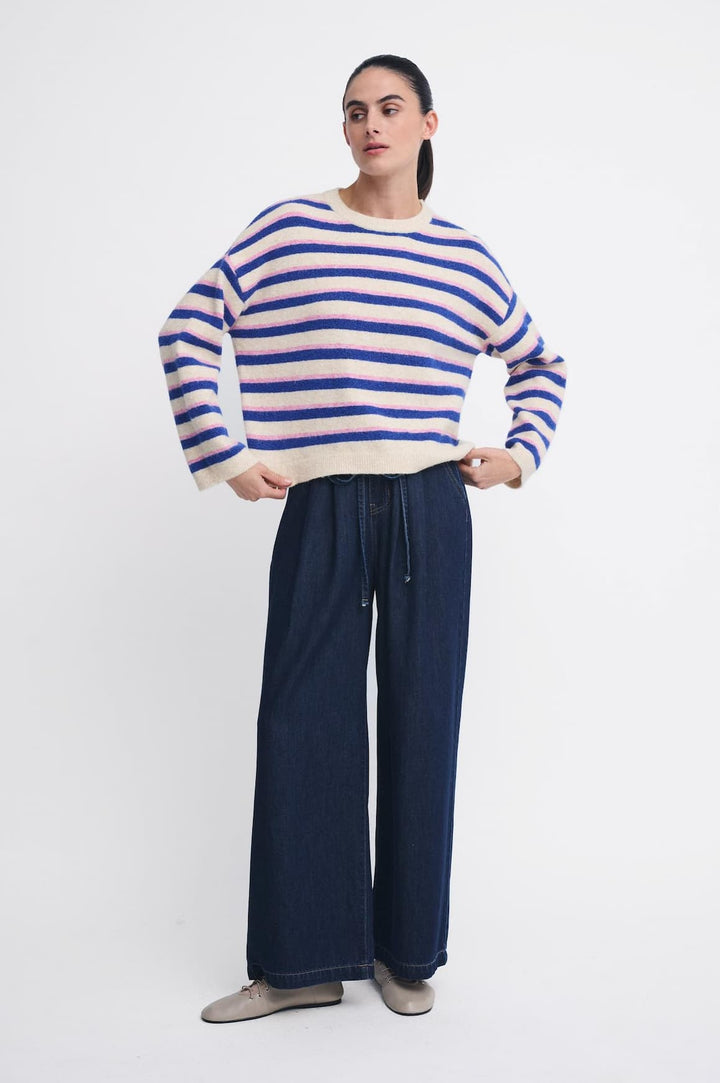 A woman modeling a cream knit sweater with bold blue horizontal stripes, paired with dark wide-leg pants and gray slip-on shoes, against a plain white studio background.