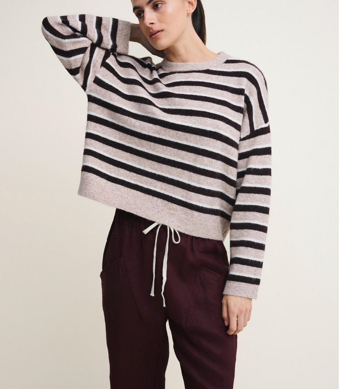 A woman wearing a cropped knit sweater with horizontal black and light beige stripes, paired with dark burgundy drawstring pants, against a plain light beige studio background.