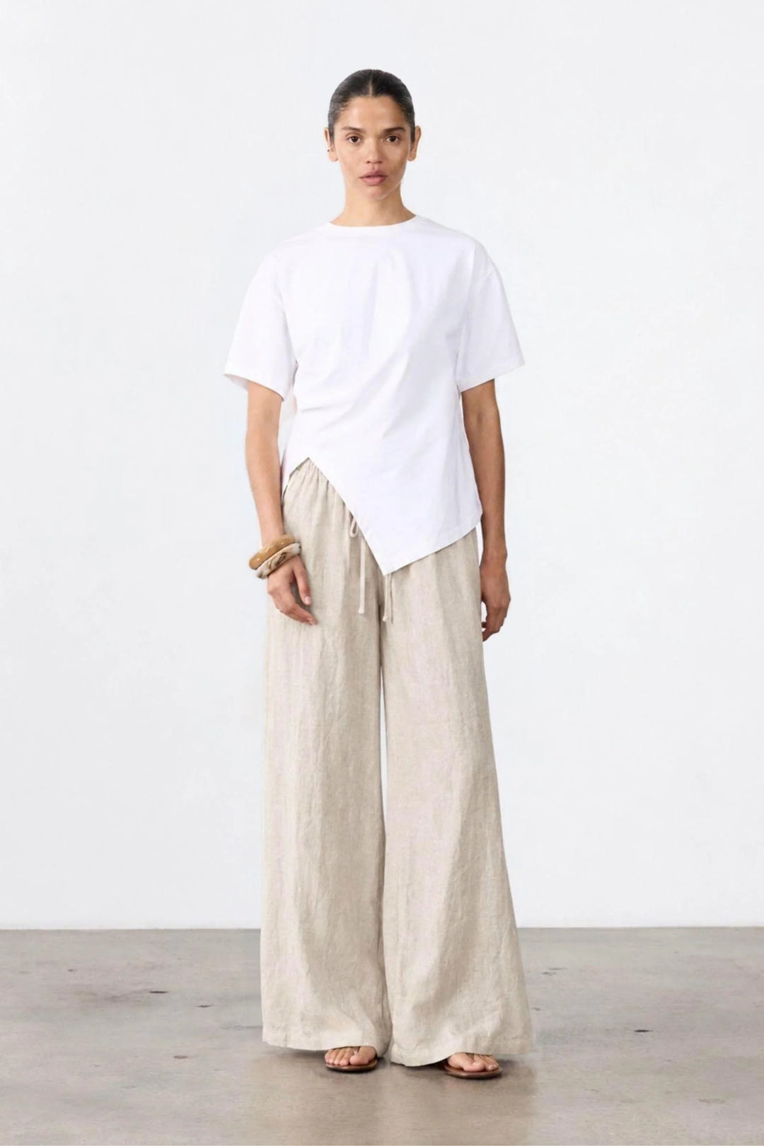 A model stands in a minimalist studio setting wearing a white asymmetrical short-sleeve t-shirt and light beige wide-leg linen trousers.