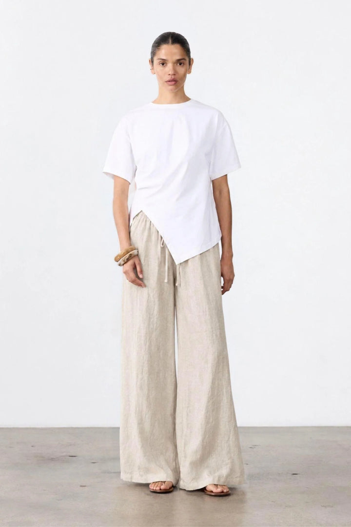 A model stands in a minimalist studio setting wearing a white asymmetrical short-sleeve t-shirt and light beige wide-leg linen trousers.