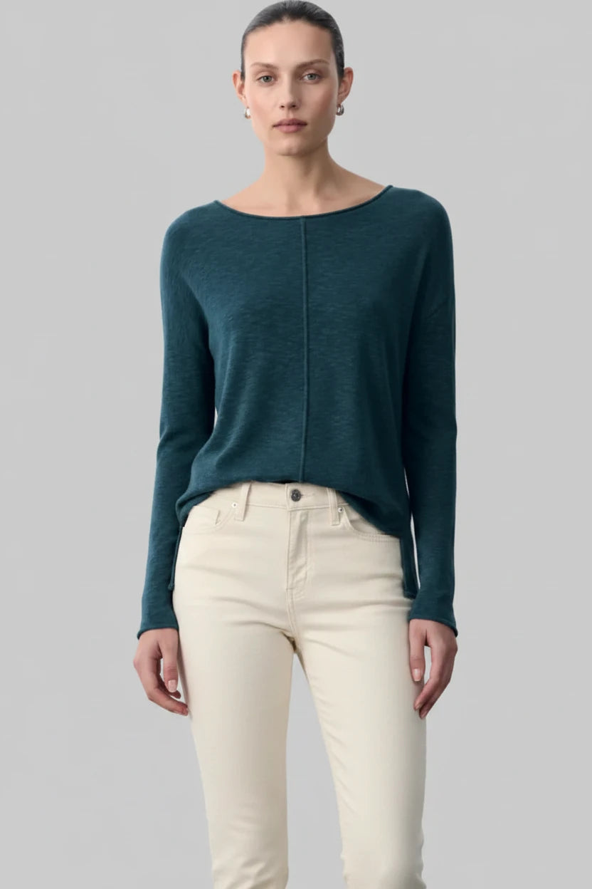 A model wearing a dark teal long-sleeve knit top with a unique front seam detail and black wide-leg trousers, shot against a solid gray background.