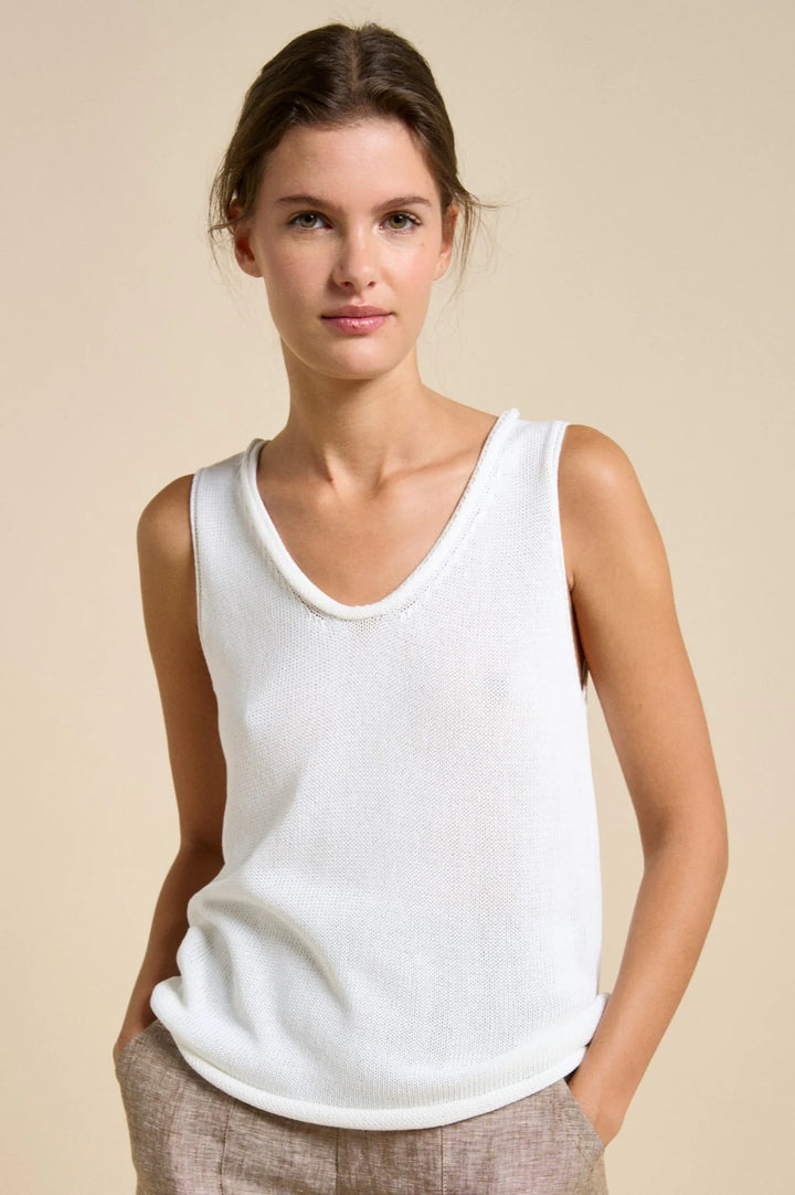 A woman wearing a white ribbed knit sleeveless tank top and light beige trousers, standing against a solid beige studio background.