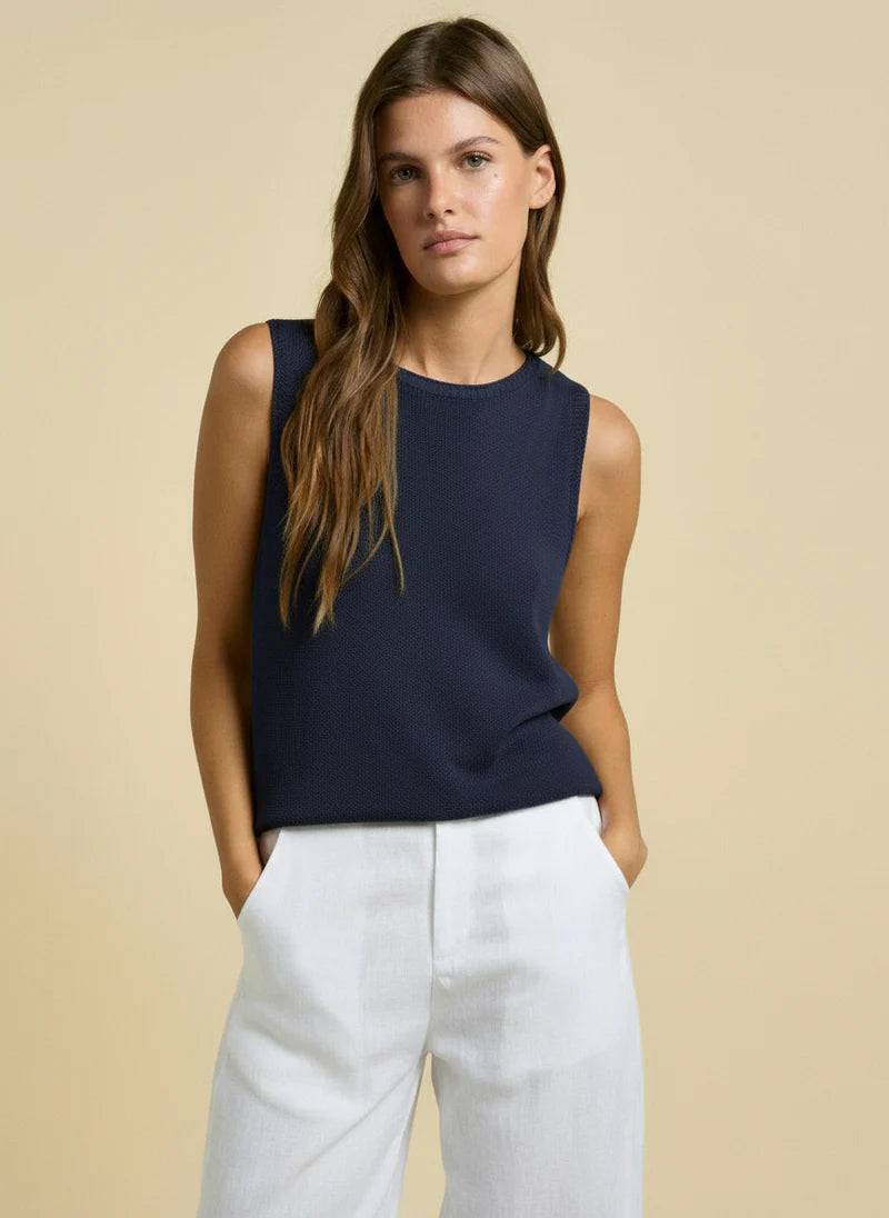 Woman wearing a navy sleeveless top and white pants against a beige background