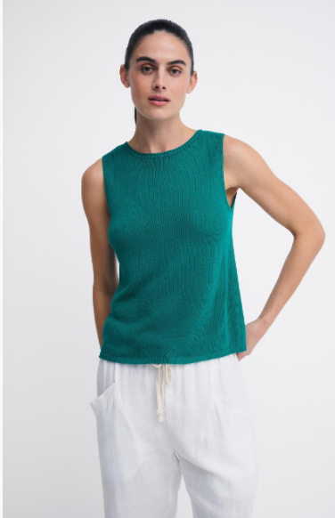 A woman modeling a sleeveless ribbed knit top in emerald green, paired with white drawstring pants, against a plain light gray studio background.