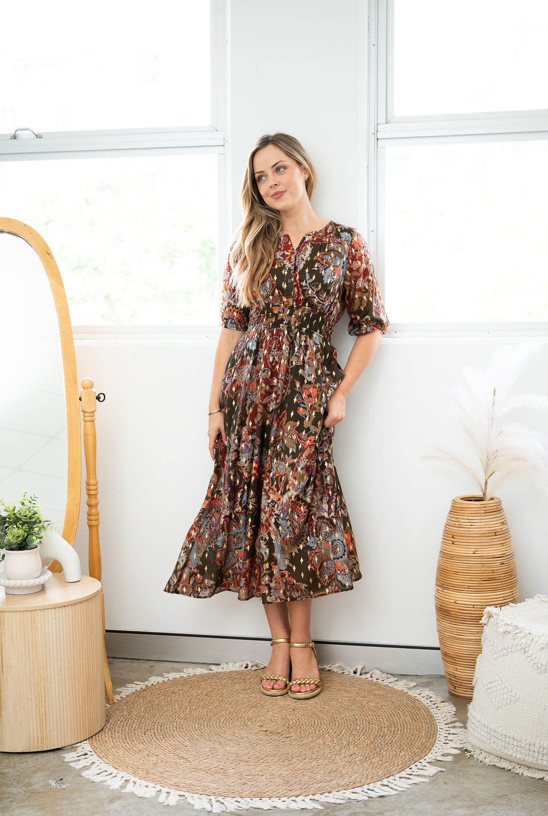 Woman in a floral dress standing in a room with a white wall and wooden furniture.