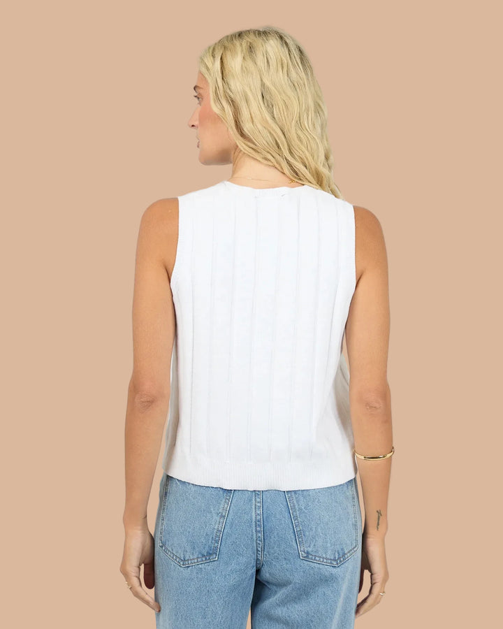 A woman shown from the back wearing a white sleeveless knit top with vertical pleats and light blue jeans, against a plain light gray studio background.