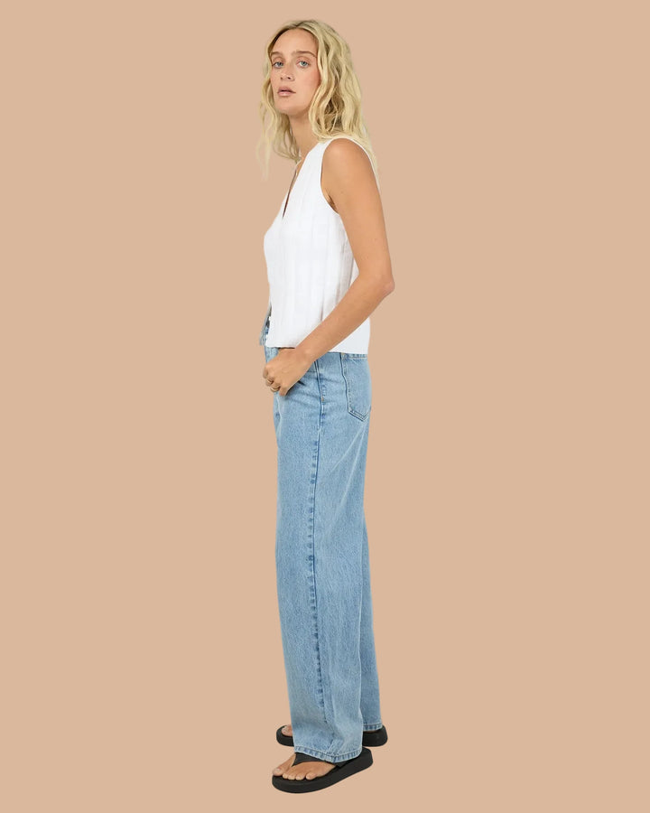 A blonde woman wearing a sleeveless white V-neck top and light blue jeans, standing against a light gray studio background.