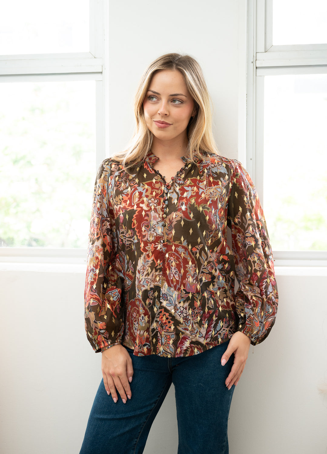 Woman wearing a patterned blouse and jeans standing in a bright room with large windows.