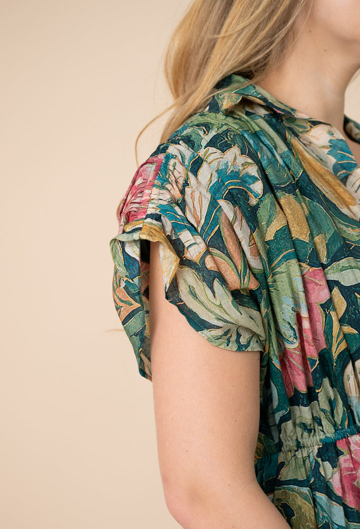 Close-up of a woman wearing a short-sleeve blouse with a tropical floral print in green, pink, beige, and brown tones against a plain beige background.