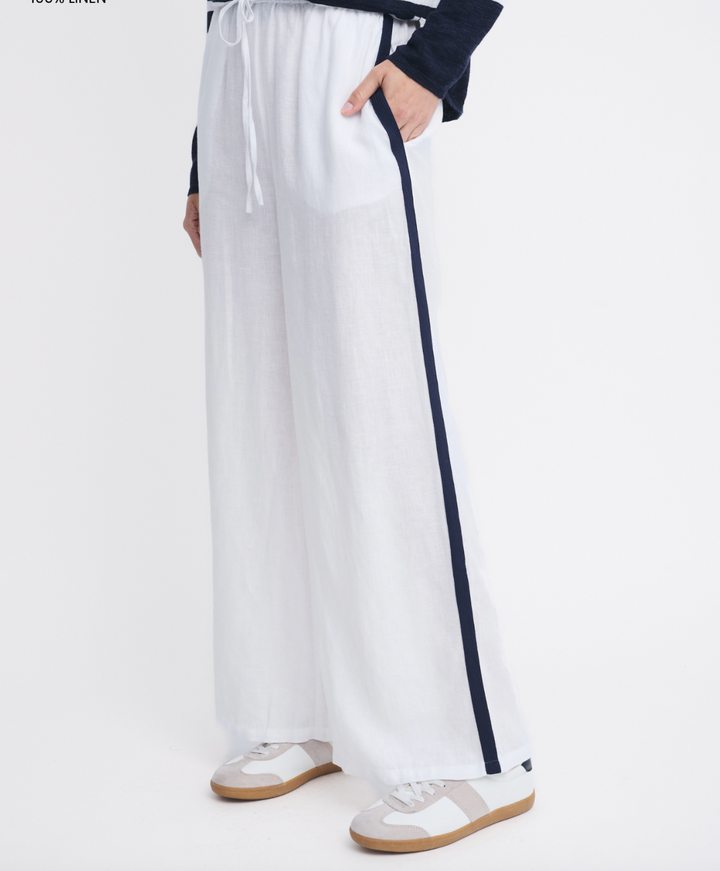White wide-leg linen pants with a navy blue side stripe, worn by a model with a navy long-sleeve top and beige sneakers against a white studio background.