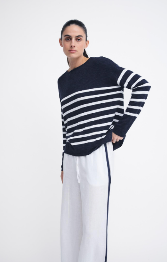 A woman modeling a navy blue and white horizontally striped knit sweater with white wide-leg pants against a plain white studio background.