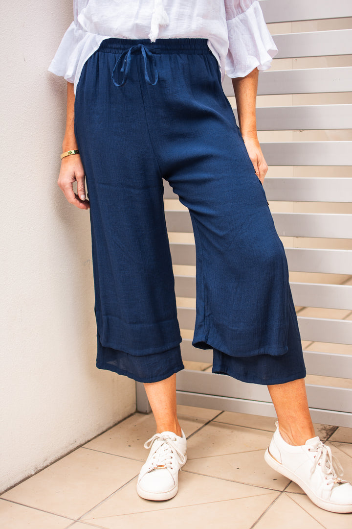 A person standing with one foot on a step wearing navy blue double-layered beach pants with an elasticized and drawstring waist, paired with a white top and white sneakers.