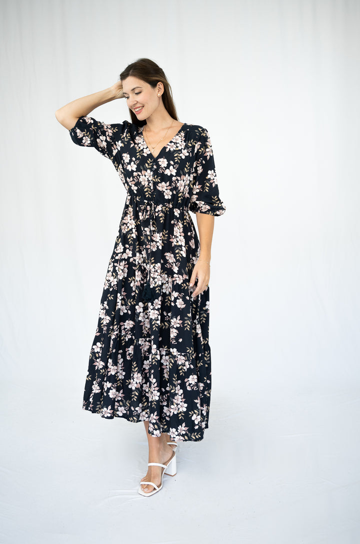 A woman modeling a black midi wrap dress with a white and pink floral pattern, featuring short flutter sleeves and a V-neckline, paired with white heeled sandals against a white studio background.