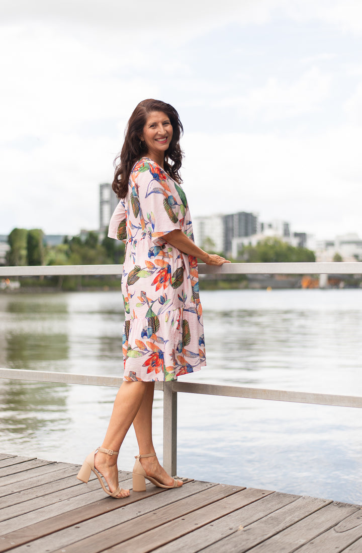 Woman in a floral dress standing on a dock by a body of water with buildings in the background.