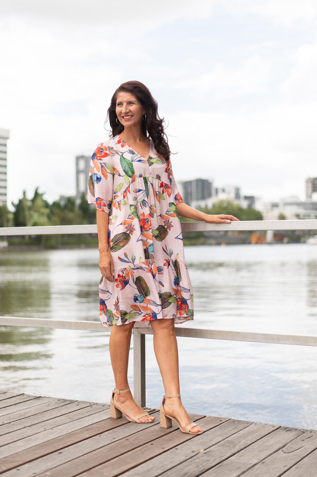Woman in a floral dress standing on a wooden dock by a body of water with buildings in the background.