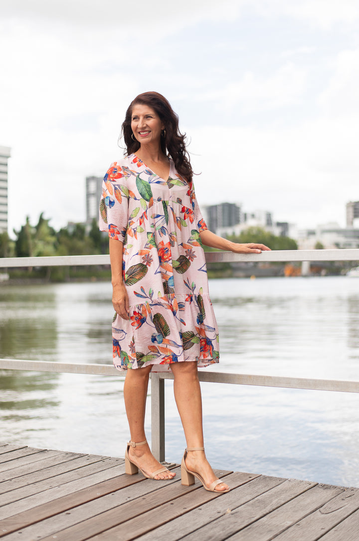Woman in a floral dress standing on a wooden dock by a body of water with buildings in the background.