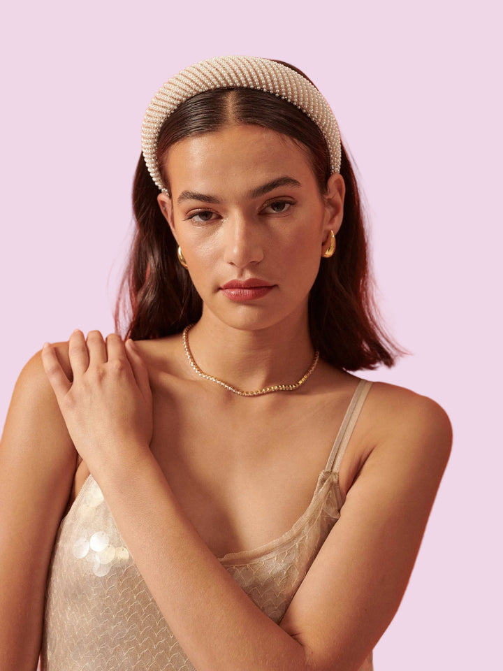 A woman wearing a textured cream beaded headband, a delicate gold chain necklace, and gold hoop earrings, paired with a champagne-colored sequined camisole against a solid light purple background.