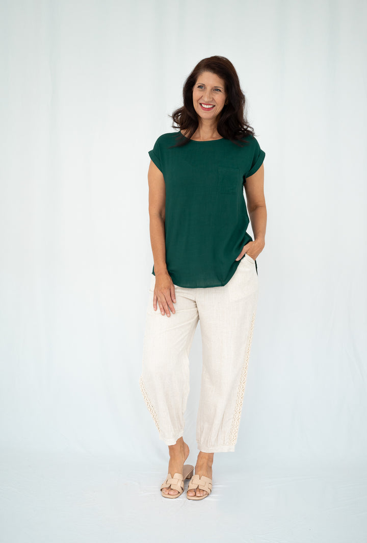 A woman modeling a dark green short-sleeve top and cream wide-leg pants against a light gray studio backdrop.