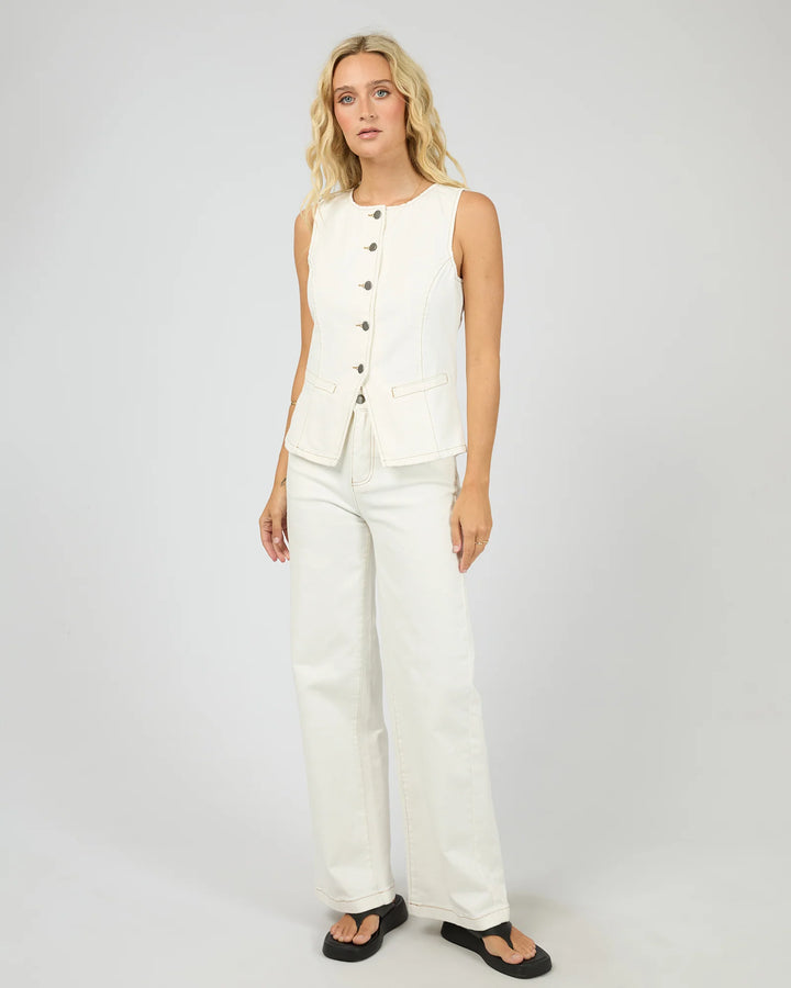 A blonde model stands against a light gray studio background wearing a sleeveless white vest with dark buttons and matching white wide-leg pants, paired with black sandals.