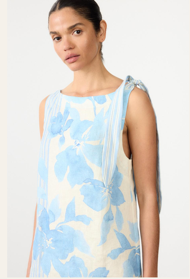 A woman wearing a sleeveless cream-colored dress with a light blue floral print and tie detail on the shoulder, photographed against a plain light background.
