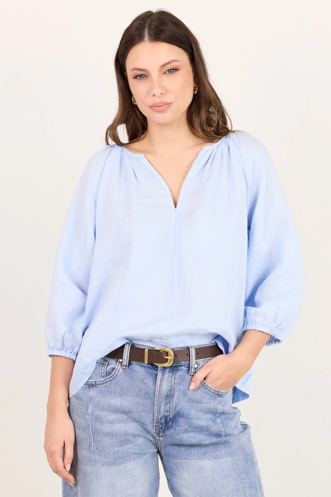 A woman wearing a light blue, loose-fitting blouse with a deep V-neck and voluminous elbow-length sleeves, paired with light-wash blue jeans and a brown belt, against a plain off-white background.