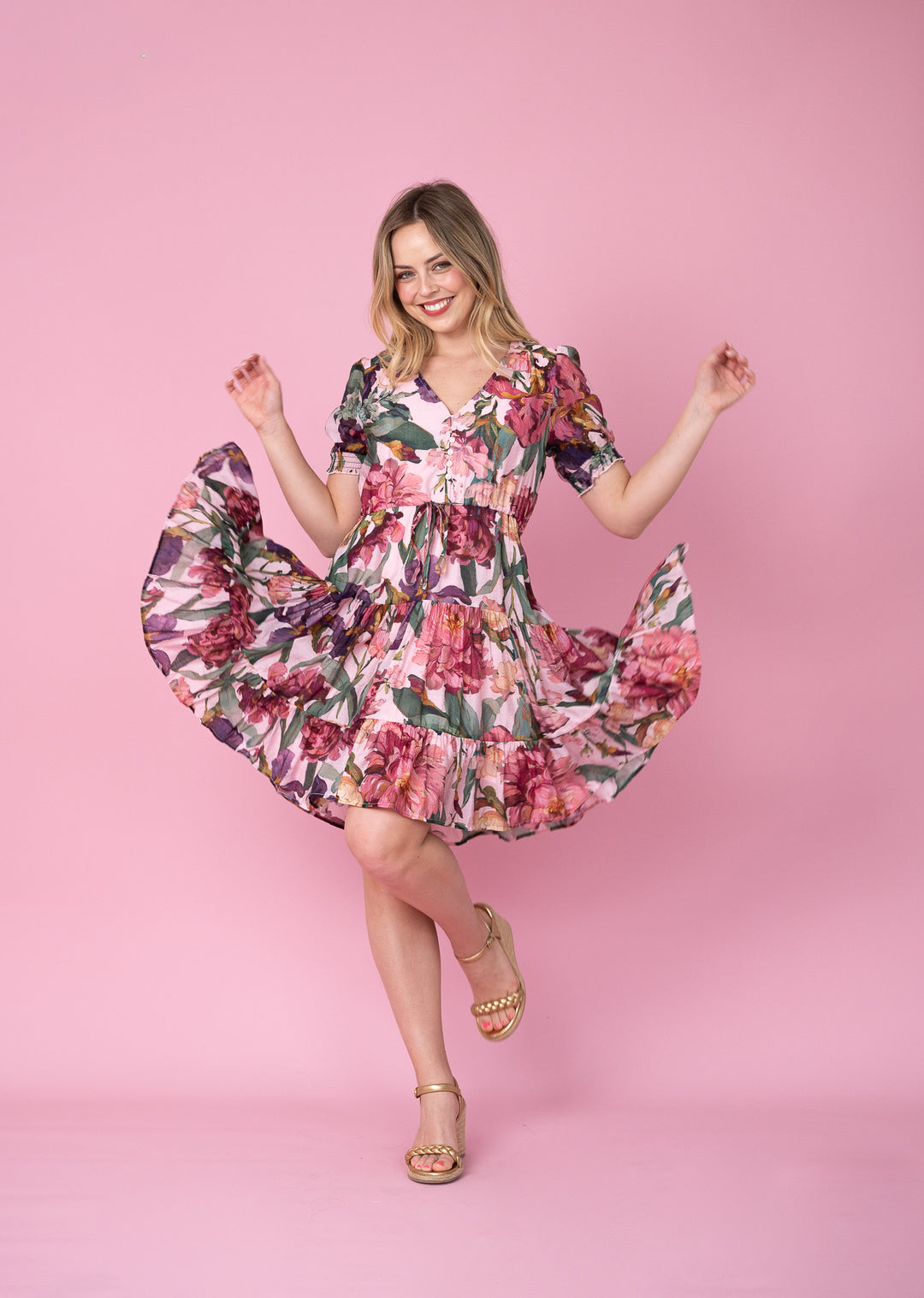 A woman posing in a pink floral dress with gold sandals against a solid pink studio background.