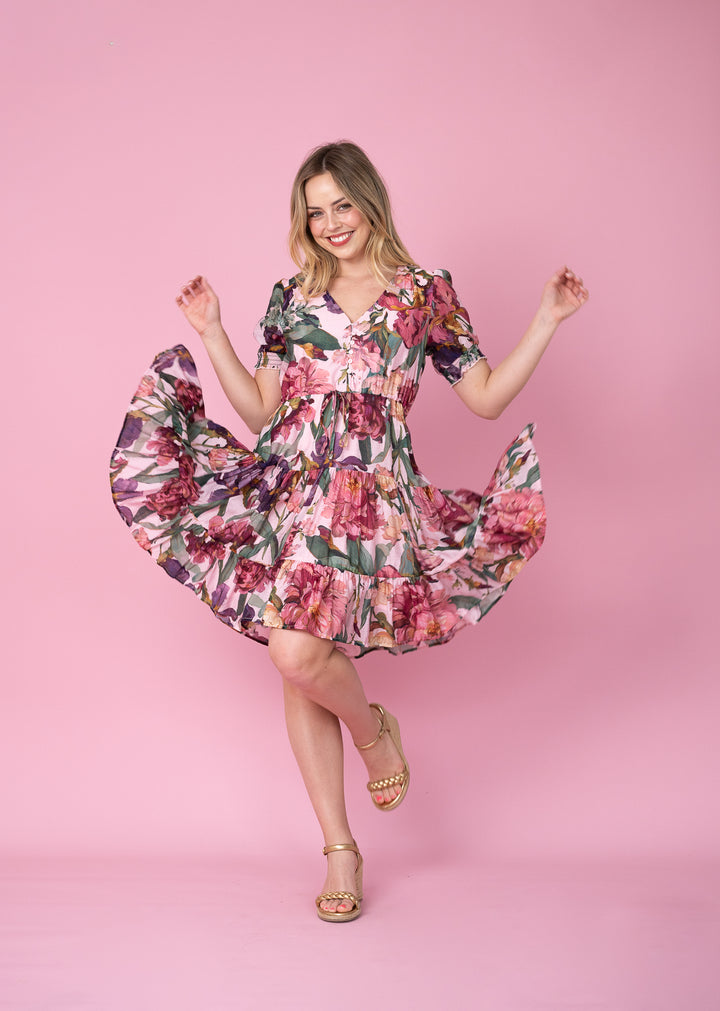 A woman posing in a pink floral dress with gold sandals against a solid pink studio background.