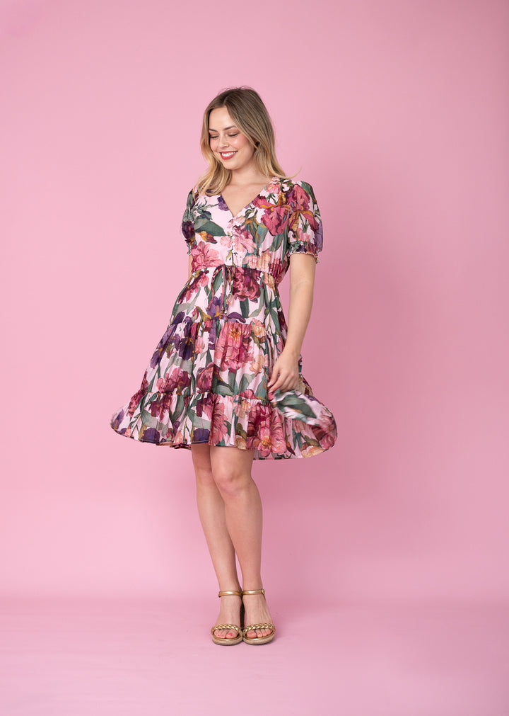 A woman modeling a short puff-sleeve V-neck dress with a pink, red, and green floral print, standing against a solid pink studio backdrop.