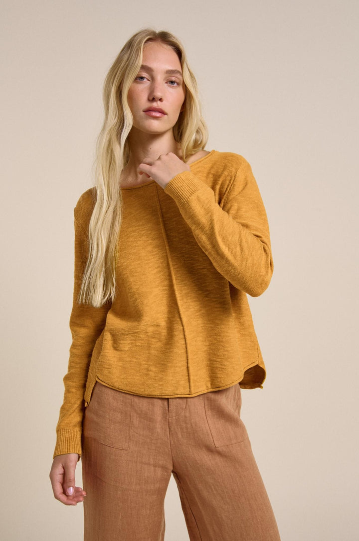 Woman wearing a mustard yellow sweater and brown pants against a beige background