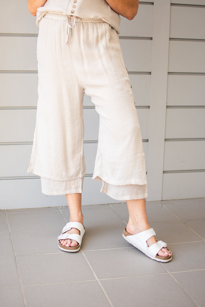 A person standing wearing beige beach pants with a drawstring waist, 7/8 length, paired with a gray top and white sandals.