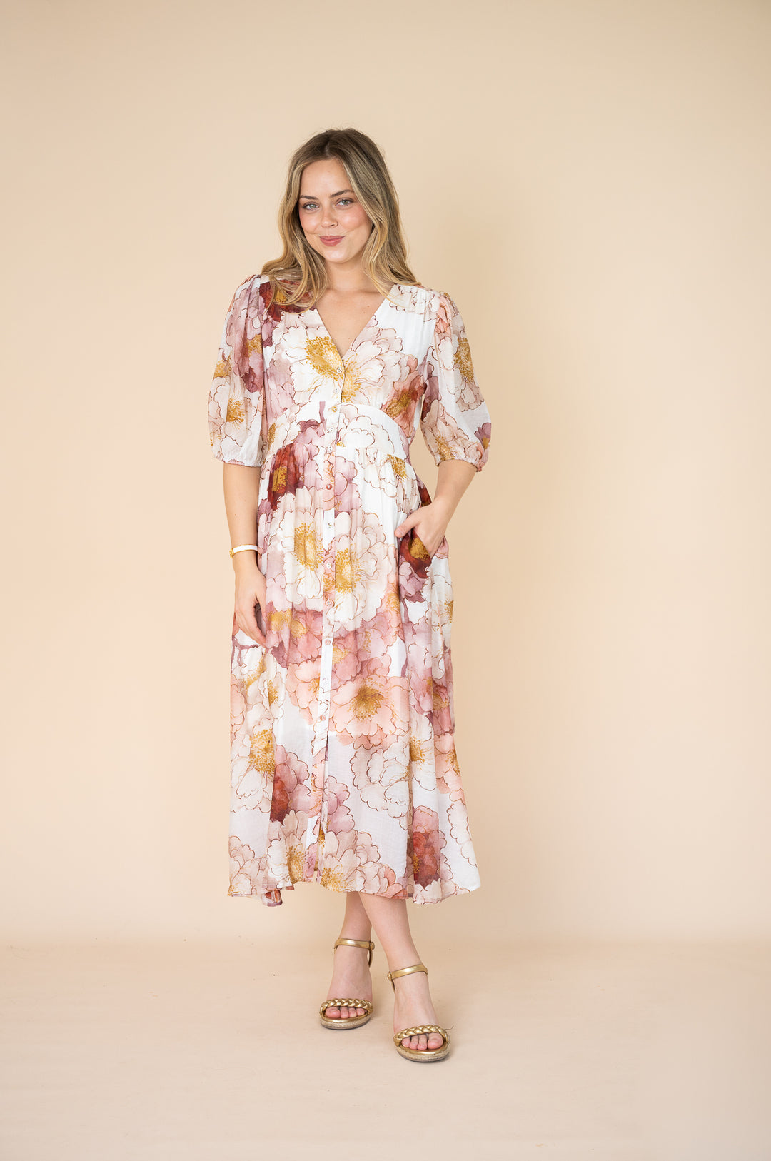 A woman modeling a long-sleeve midi wrap dress with a watercolor floral print in pink, orange, and yellow on a white background, set against a solid beige backdrop.