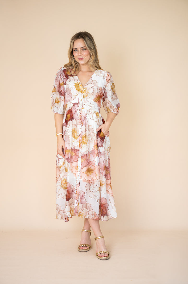 A woman modeling a long-sleeve midi wrap dress with a watercolor floral print in pink, orange, and yellow on a white background, set against a solid beige backdrop.