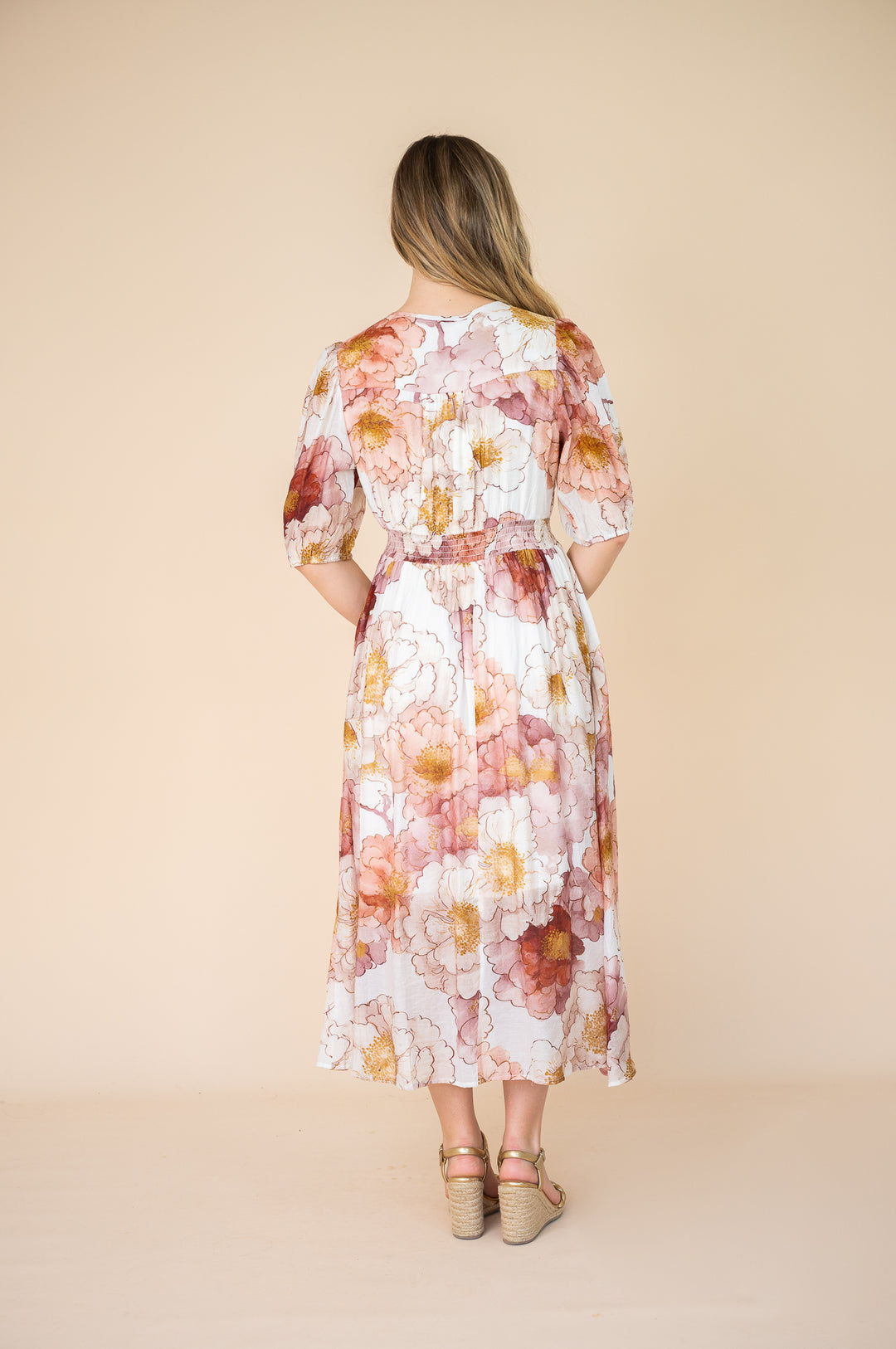 A woman viewed from behind wearing a midi-length floral dress with puff sleeves and a tie waist, set against a solid beige studio background.