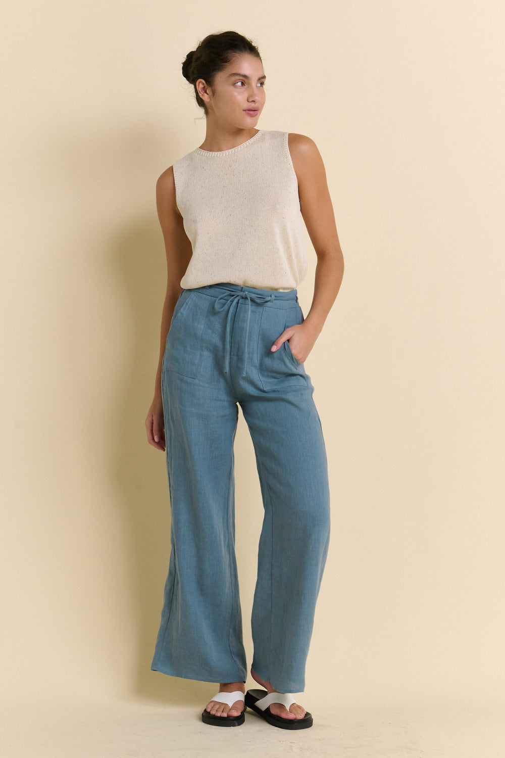 Woman wearing a sleeveless top and blue pants against a beige background